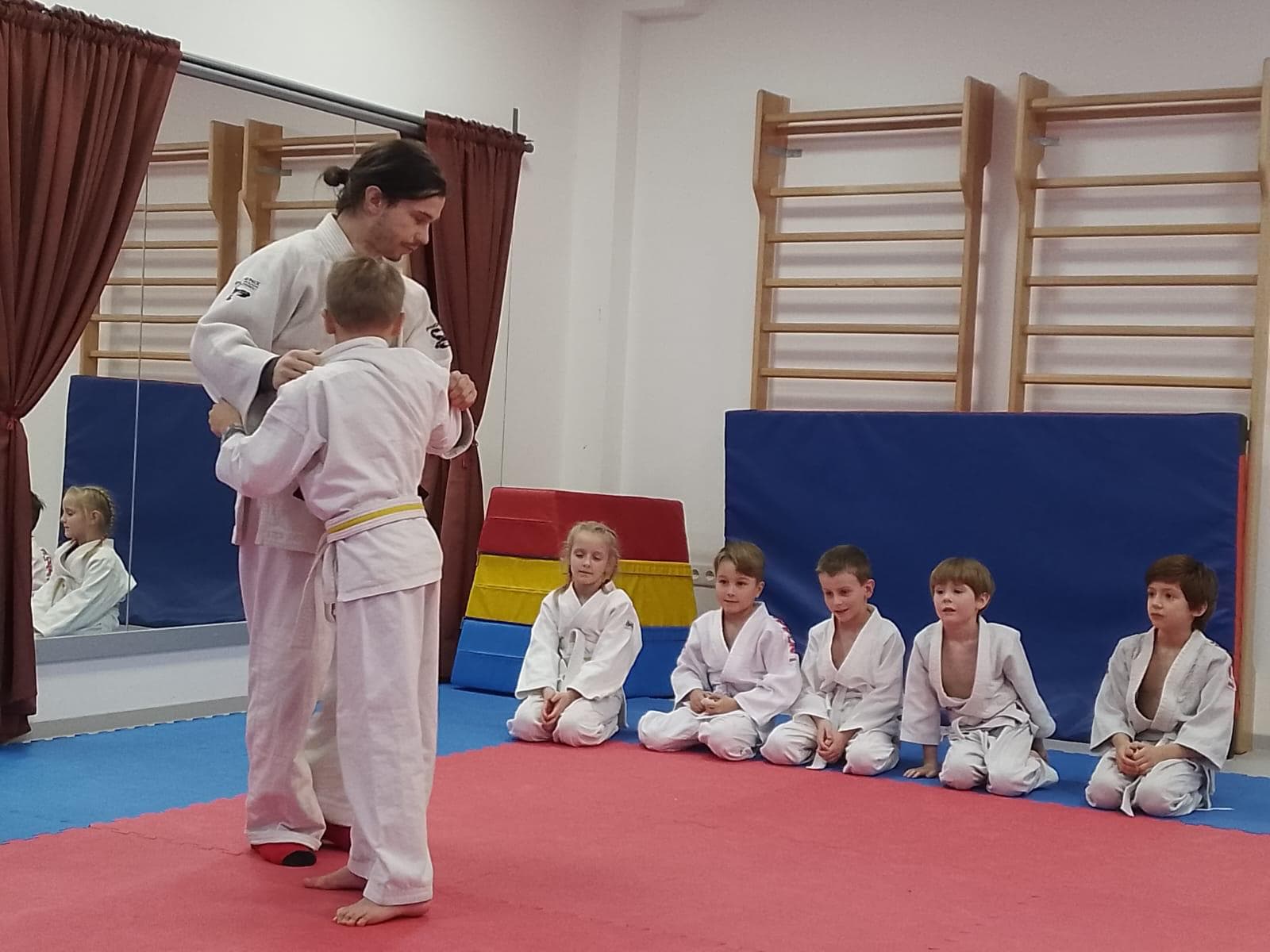 Trenér pomáhá malému žákovi s kimonem, ostatní děti přihlížejí